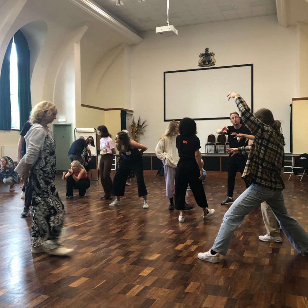 A group of people engaged in expressive movements in a large room with wooden floors, high ceilings, and a coat of arms on the wall.