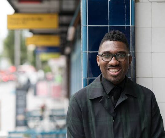 A man wearing glasses and a dark coat smiles at the camera as he stands outdoors beside a building with blue and white tiles. The background shows a blurry urban setting with signs and vehicles.