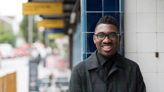 A man wearing glasses and a dark coat smiles at the camera as he stands outdoors beside a building with blue and white tiles. The background shows a blurry urban setting with signs and vehicles.