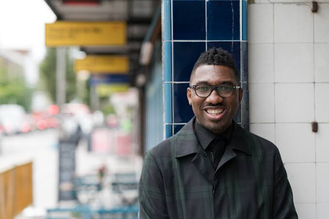 A man wearing glasses and a dark coat smiles at the camera as he stands outdoors beside a building with blue and white tiles. The background shows a blurry urban setting with signs and vehicles.