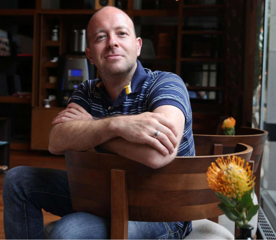 A man with a shaved head and striped shirt sits in a modern room, leaning on the back of a chair with arms crossed. A small orange flower is visible in the foreground.