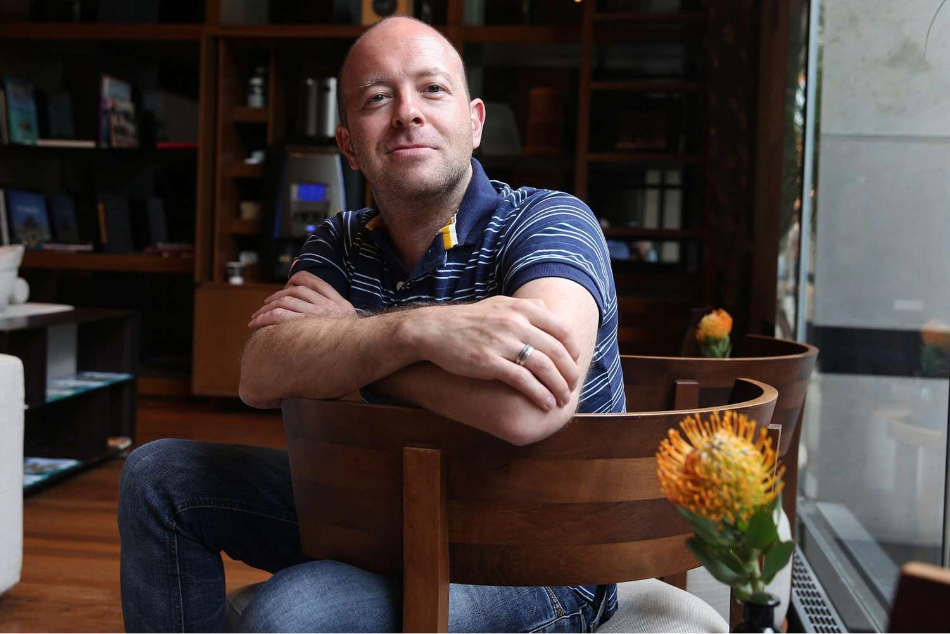 A man with a shaved head and striped shirt sits in a modern room, leaning on the back of a chair with arms crossed. A small orange flower is visible in the foreground.