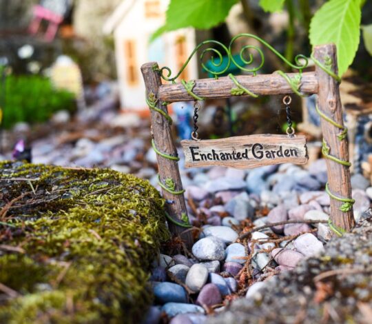 A wooden sign reading "Enchanted Garden" hangs on a small wooden gate surrounded by pebbles and moss, with a miniature house and plants in the background.