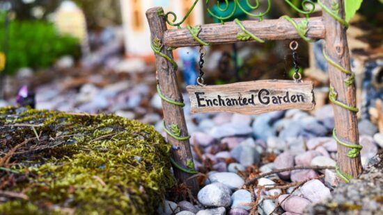 A wooden sign reading "Enchanted Garden" hangs on a small wooden gate surrounded by pebbles and moss, with a miniature house and plants in the background.