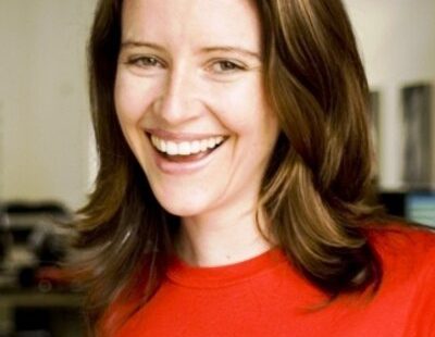 A woman with shoulder-length brown hair wearing a red shirt smiles brightly in an indoor setting.