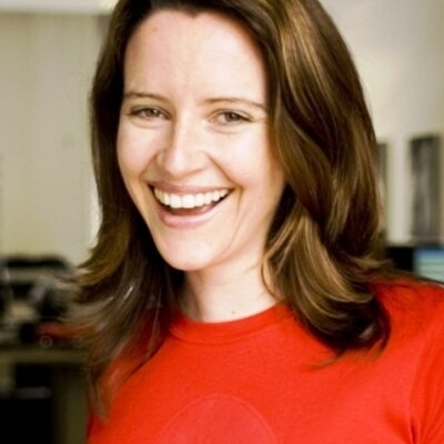 A woman with shoulder-length brown hair wearing a red shirt smiles brightly in an indoor setting.