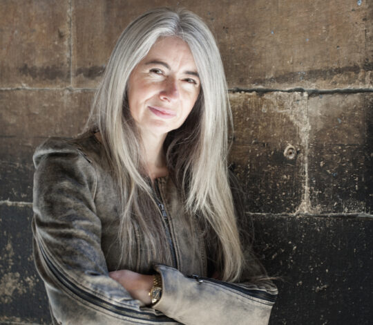 A person with long, gray hair is standing against a weathered stone wall, wearing a brown jacket, arms crossed, and smiling slightly.