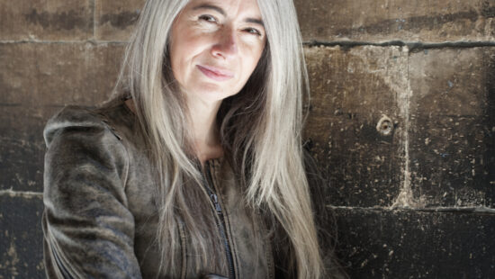 A person with long, gray hair is standing against a weathered stone wall, wearing a brown jacket, arms crossed, and smiling slightly.