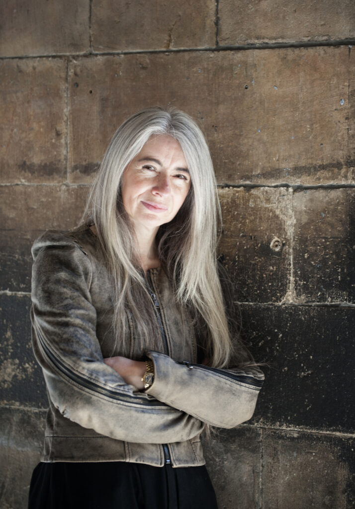 A person with long, gray hair is standing against a weathered stone wall, wearing a brown jacket, arms crossed, and smiling slightly.