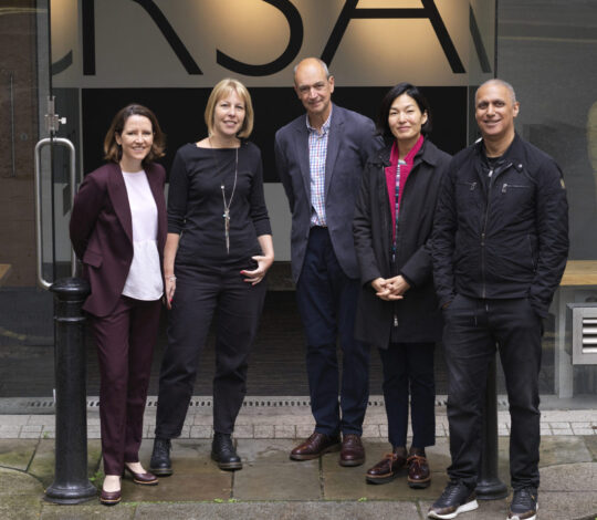 A group of five adults, three women and two men, standing outside a building with large windows displaying the letters "RSA". They are dressed in business-casual attire and smiling at the camera.