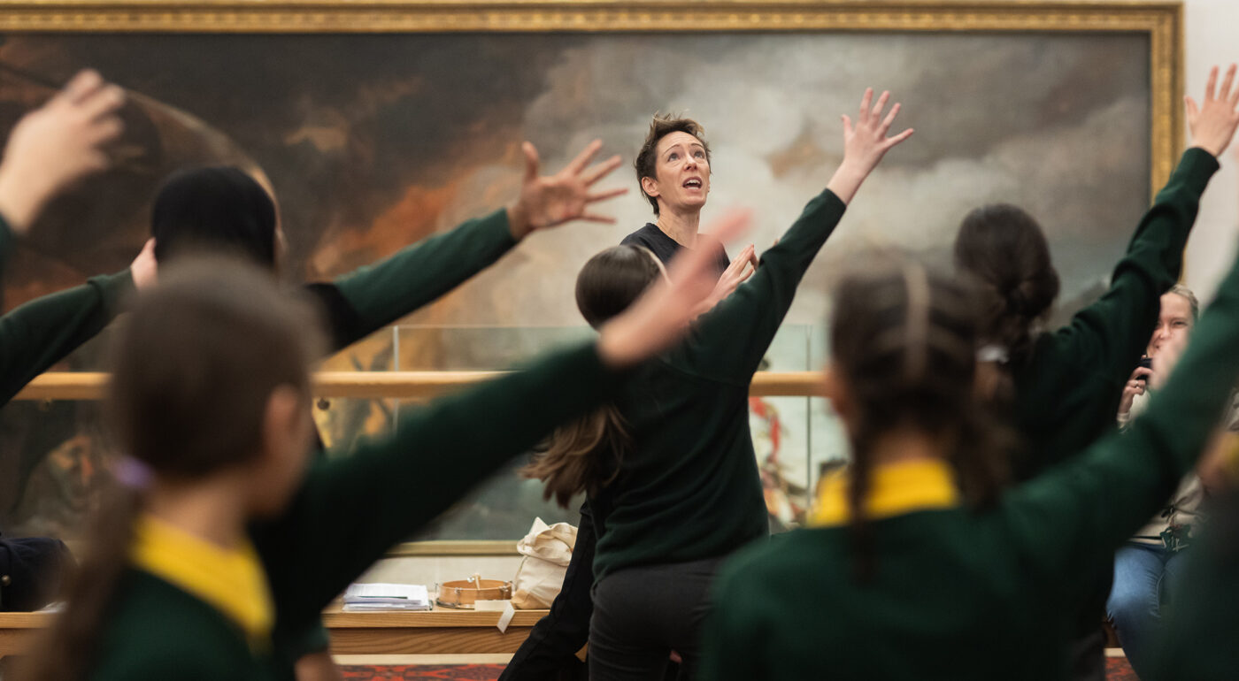 A teacher leads a group of students in a classroom activity, all raising their arms. A large painting hangs on the wall in the background.