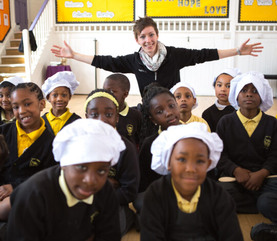 A woman stands with arms outstretched behind a group of school children. Some children wear white chef hats, and they are all seated in a classroom. Yellow bulletin boards are in the background.