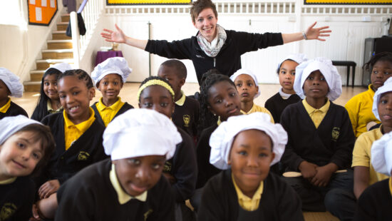 A woman stands with arms outstretched behind a group of school children. Some children wear white chef hats, and they are all seated in a classroom. Yellow bulletin boards are in the background.