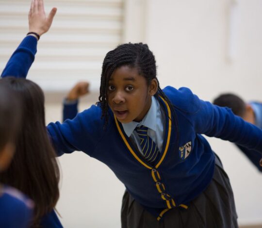 A student in a blue school uniform and tie participates in a classroom activity, extending her arm while other students are engaged in the background.