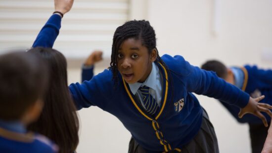 A student in a blue school uniform and tie participates in a classroom activity, extending her arm while other students are engaged in the background.