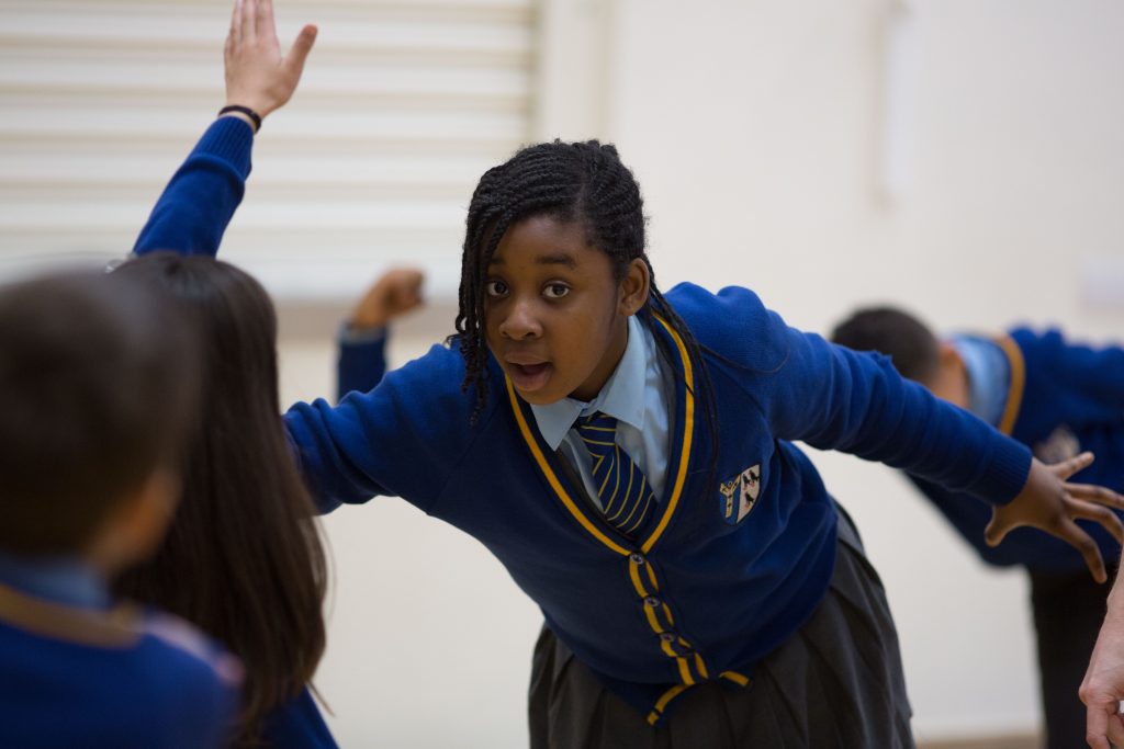 A student in a blue school uniform and tie participates in a classroom activity, extending her arm while other students are engaged in the background.