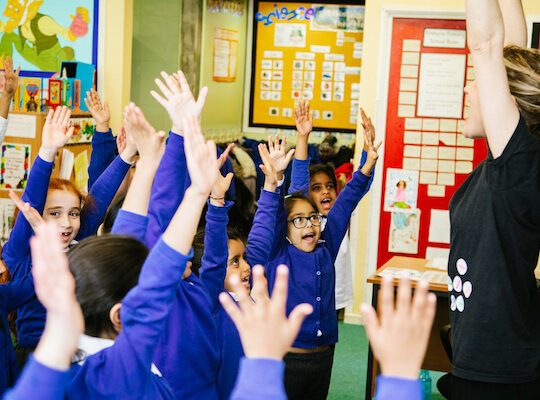 A group of children in blue uniforms raise their hands in a classroom while a teacher with raised arms leads them. Bright artwork and educational posters are visible on the walls.