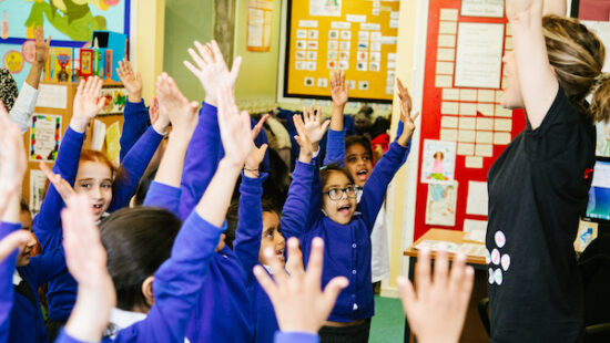 A group of children in blue uniforms raise their hands in a classroom while a teacher with raised arms leads them. Bright artwork and educational posters are visible on the walls.