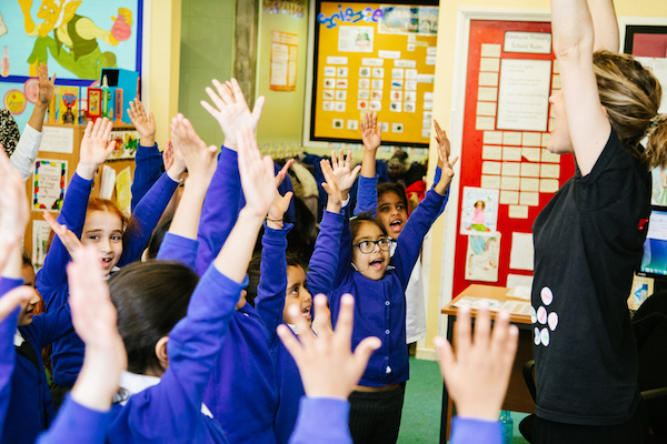 A group of children in blue uniforms raise their hands in a classroom while a teacher with raised arms leads them. Bright artwork and educational posters are visible on the walls.