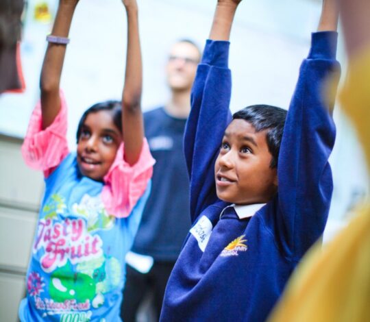 Two children stand with their arms raised, one wearing a colorful shirt and the other in a blue school uniform, smiling and looking up.