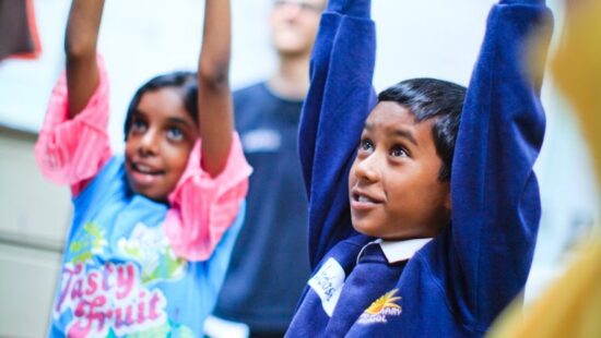 Two children stand with their arms raised, one wearing a colorful shirt and the other in a blue school uniform, smiling and looking up.