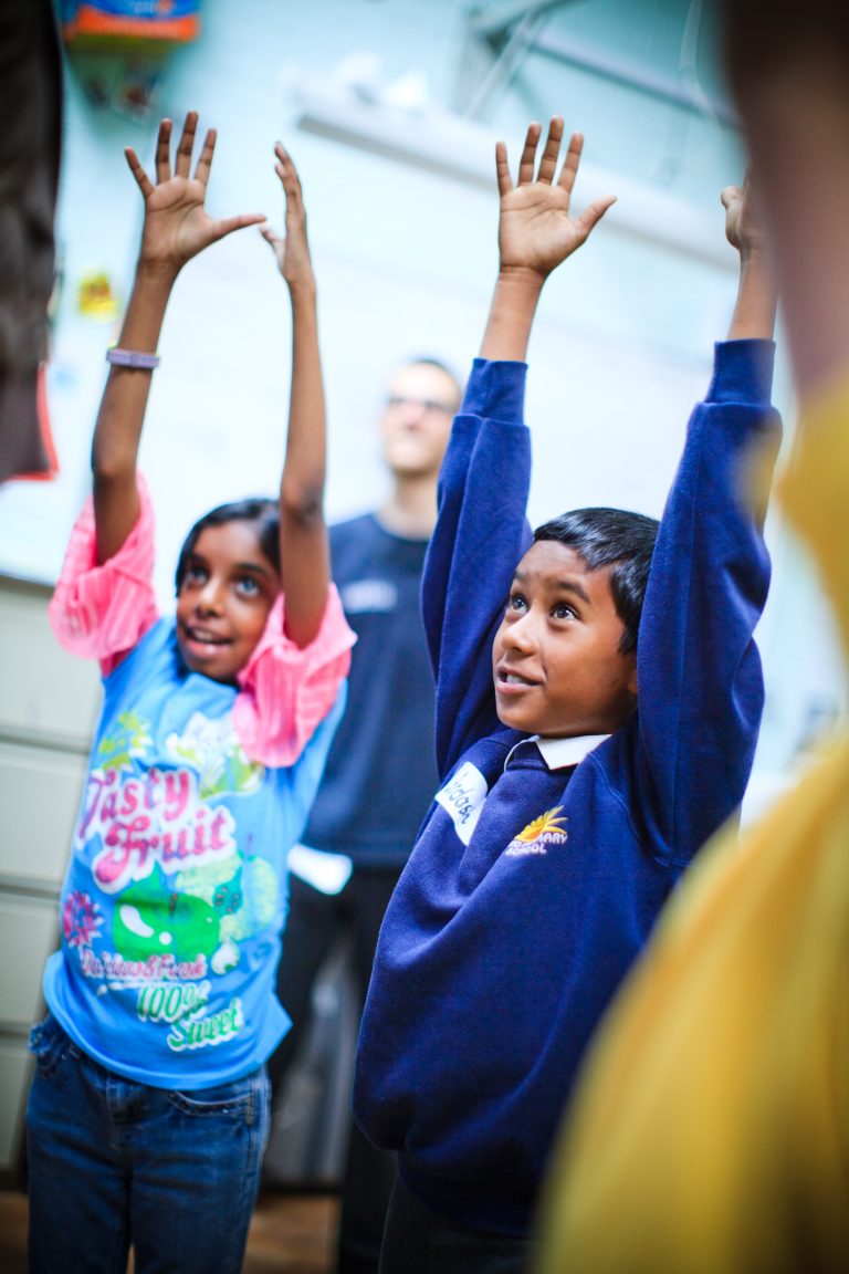 Two children stand with their arms raised, one wearing a colorful shirt and the other in a blue school uniform, smiling and looking up.