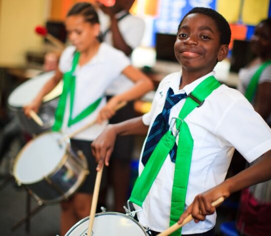 Children in school uniforms play drums, practicing in a classroom with bulletin boards in the background.