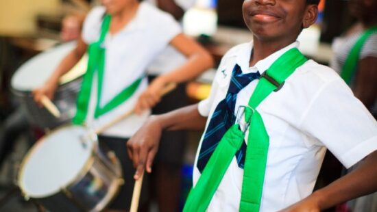 Children in school uniforms play drums, practicing in a classroom with bulletin boards in the background.