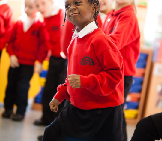 Children in uniform participate in an activity at school. The focus is on a child in a red sweater and navy skirt, smiling and walking purposefully, with other children blurred in the background.