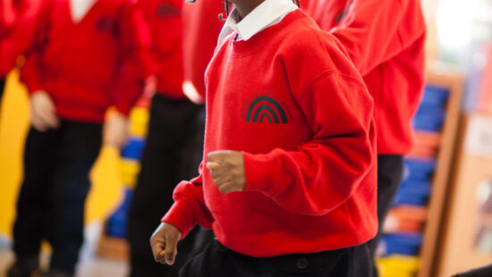 Children in uniform participate in an activity at school. The focus is on a child in a red sweater and navy skirt, smiling and walking purposefully, with other children blurred in the background.