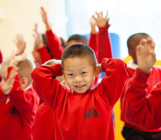 Children in a classroom wearing matching red uniforms, raising their hands and smiling. One child in the foreground is posing with their hands behind their head.