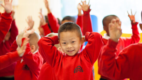 Children in a classroom wearing matching red uniforms, raising their hands and smiling. One child in the foreground is posing with their hands behind their head.