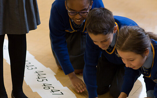 Four children in school uniforms are kneeling on the floor, looking at a number line. Another child stands nearby with only legs visible. The setting appears to be a classroom or educational environment.