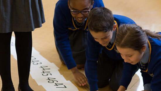 Four children in school uniforms are kneeling on the floor, looking at a number line. Another child stands nearby with only legs visible. The setting appears to be a classroom or educational environment.