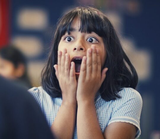 A young girl with black hair and bangs is indoors, holding her face with both hands, and expressing shock or surprise.