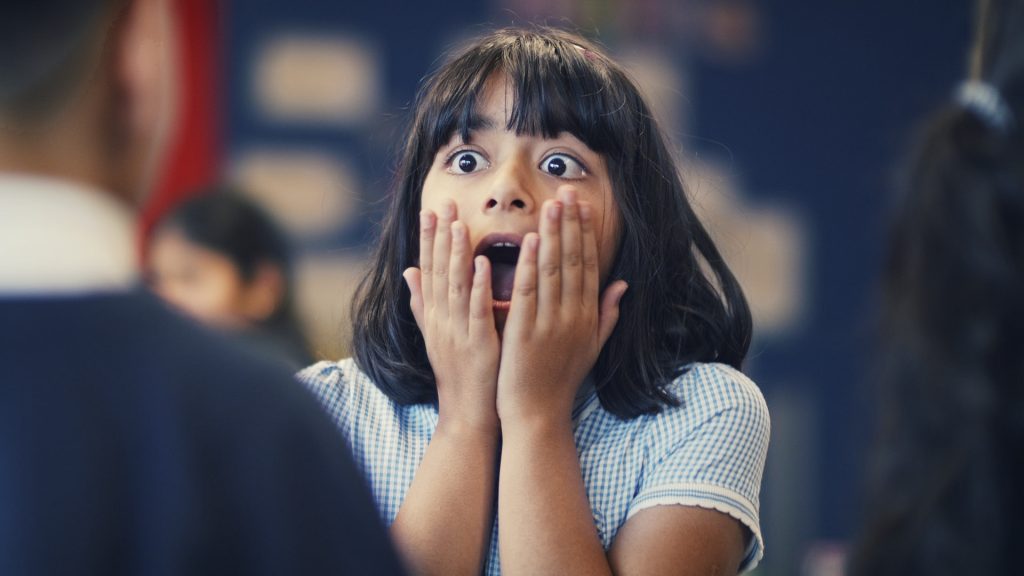 A young girl with black hair and bangs is indoors, holding her face with both hands, and expressing shock or surprise.