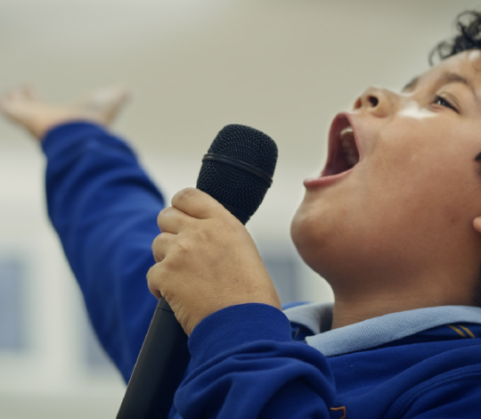 Child in a blue shirt passionately singing into a microphone with one hand raised.