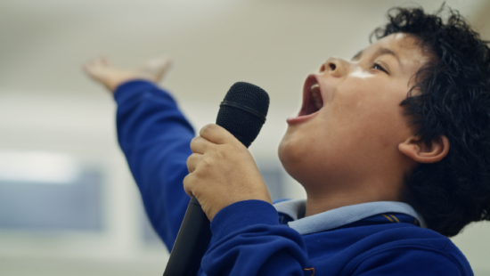 Child in a blue shirt passionately singing into a microphone with one hand raised.