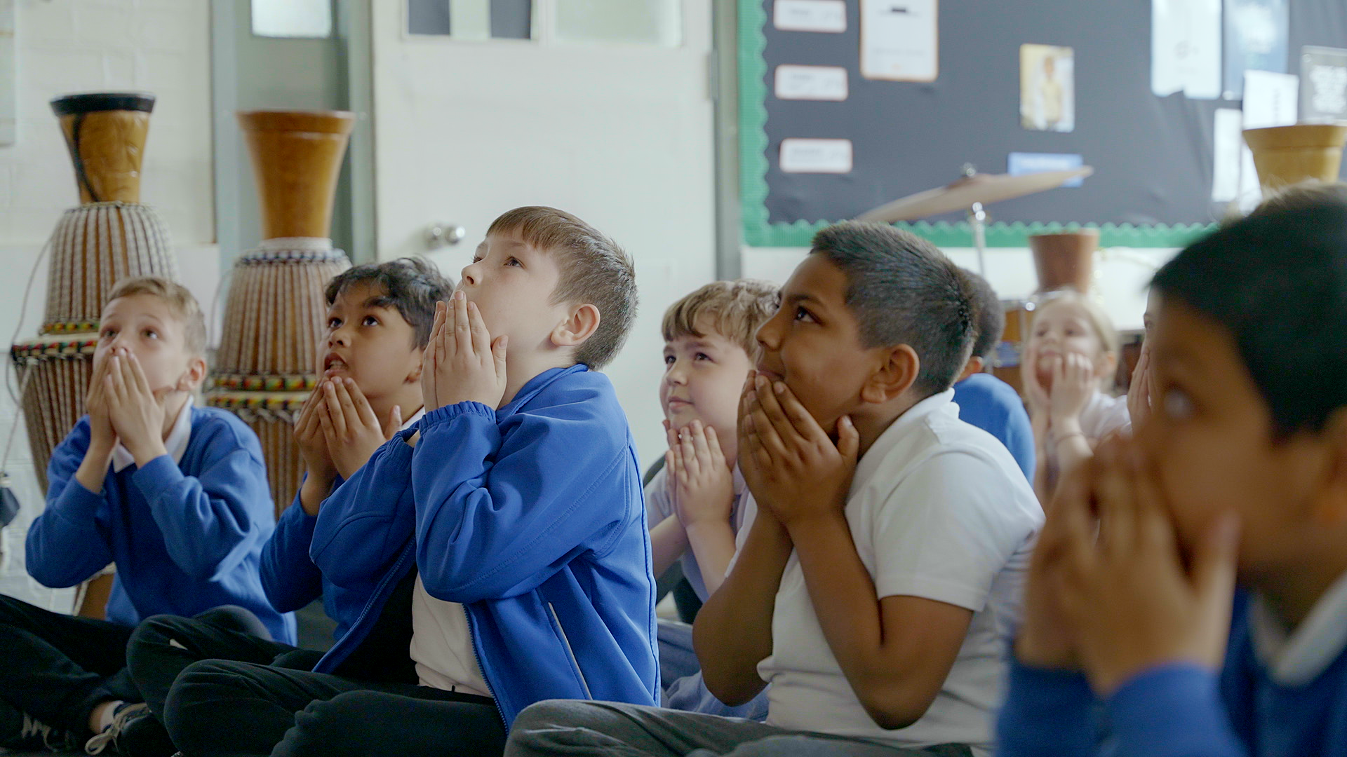 A group of children sit on the floor in a classroom with hands clasped under their chins, looking attentively towards the front. They are wearing blue and white uniforms.