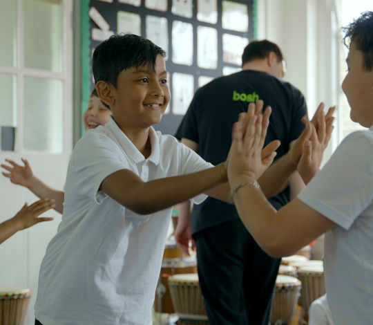 Children in a classroom playing a clapping game, wearing white shirts, with drums in the background and a person standing near the door.