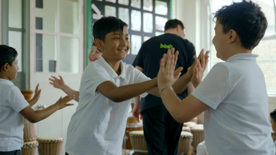 Children in a classroom playing a clapping game, wearing white shirts, with drums in the background and a person standing near the door.