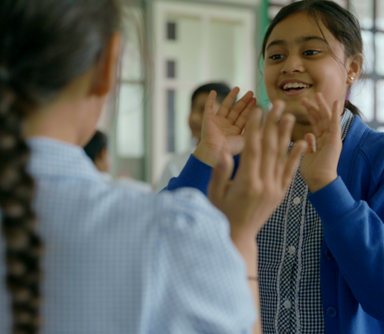 Two young girls in school uniforms are playing a hand clapping game in a classroom, smiling and enjoying their interaction.