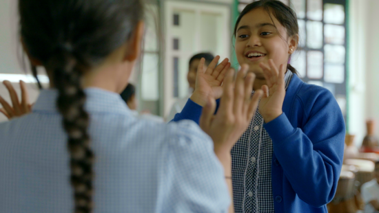 Two young girls in school uniforms are playing a hand clapping game in a classroom, smiling and enjoying their interaction.