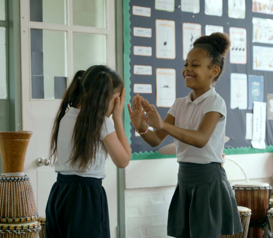 Two young girls in school uniforms are playing a clapping game in a classroom filled with traditional drums. One girl is laughing while the other covers her face with her hands. Bulletin boards are in the background.