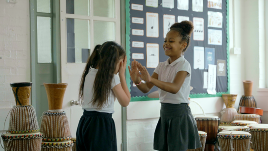 Two young girls in school uniforms are playing a clapping game in a classroom filled with traditional drums. One girl is laughing while the other covers her face with her hands. Bulletin boards are in the background.