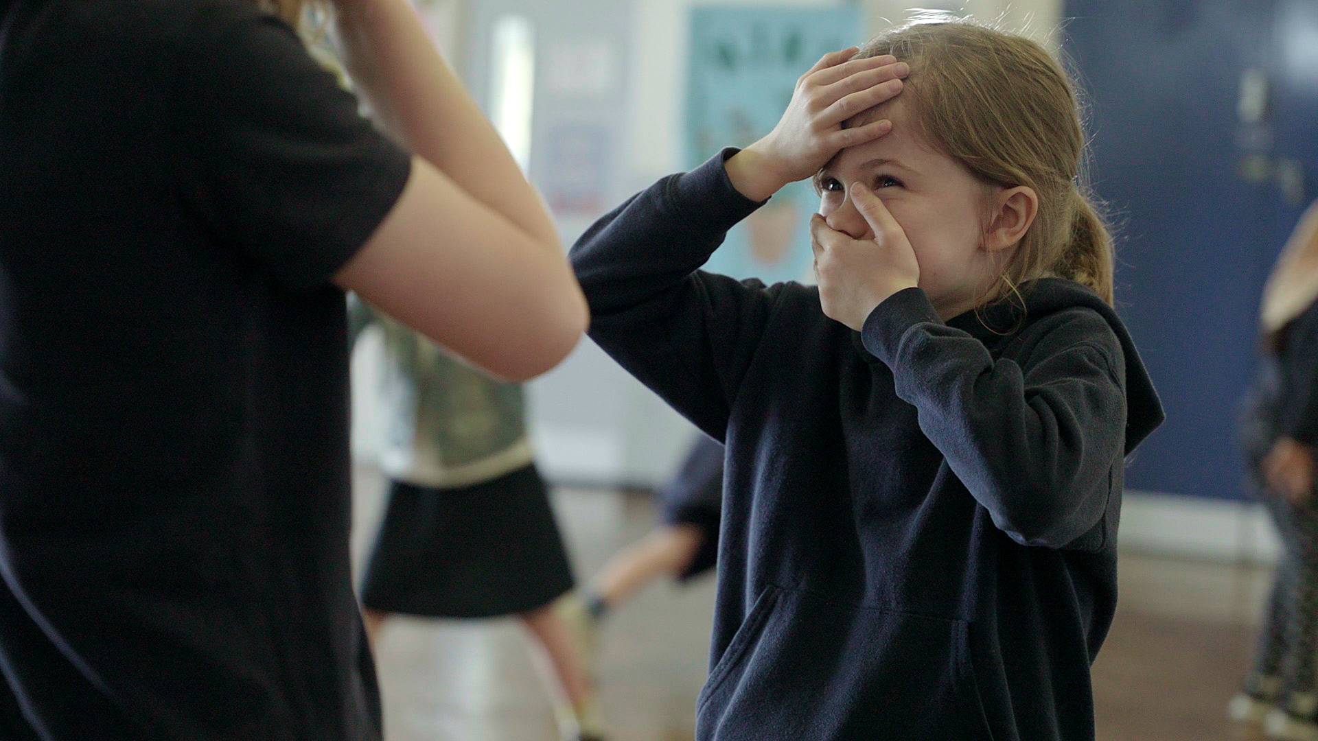 Child in a dark hoodie covers their mouth with one hand and forehead with the other, standing indoors with a blurred person in the foreground.