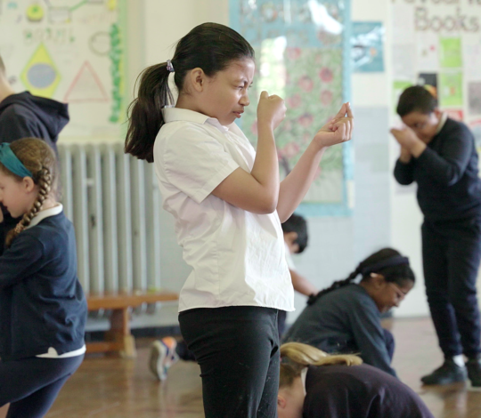 Children in a classroom engaged in an activity, with some standing and some kneeling, demonstrating focused participation.