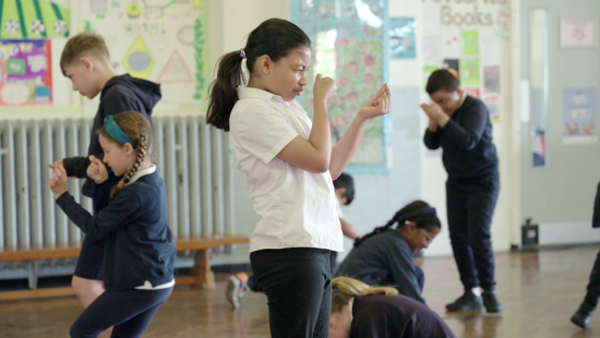 Children in a classroom engaged in an activity, with some standing and some kneeling, demonstrating focused participation.