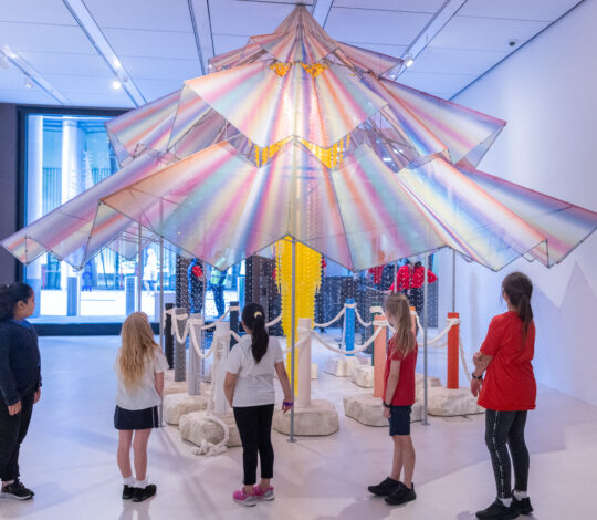 Children and adults observe a colorful, umbrella-shaped art installation in a well-lit gallery. The installation is surrounded by ropes and features various materials and textures.