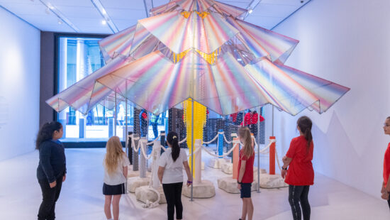 Children and adults observe a colorful, umbrella-shaped art installation in a well-lit gallery. The installation is surrounded by ropes and features various materials and textures.